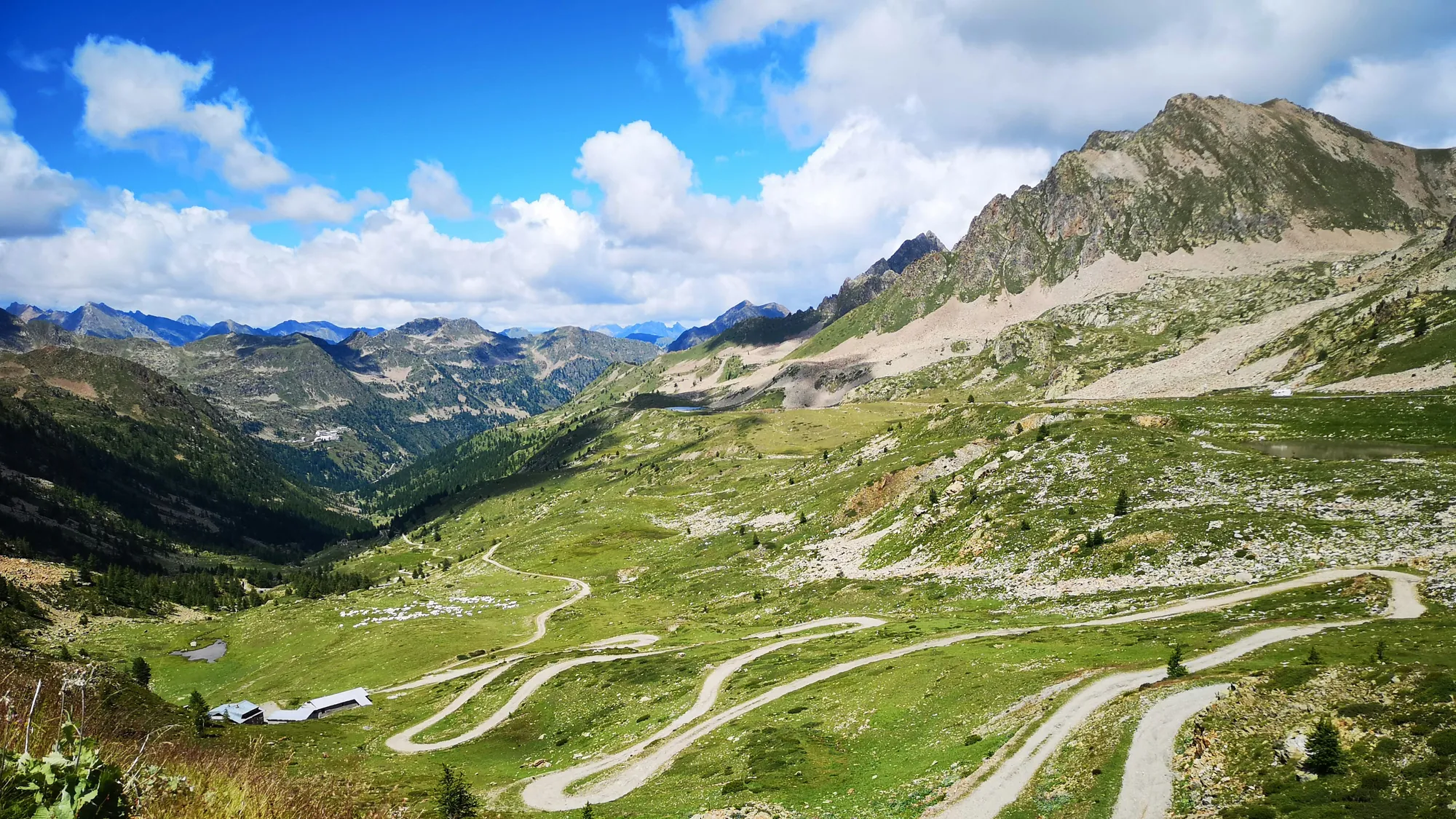 Les lacets de la route du col de la Lombarde à travers les alpages verts, sous un ciel bleu et des nuages